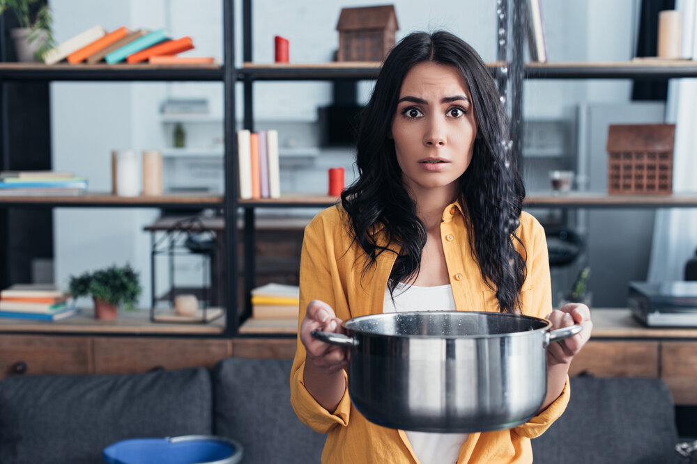 Shocked Brunette Woman Holding Pot Under Water Leaking From Ceiling