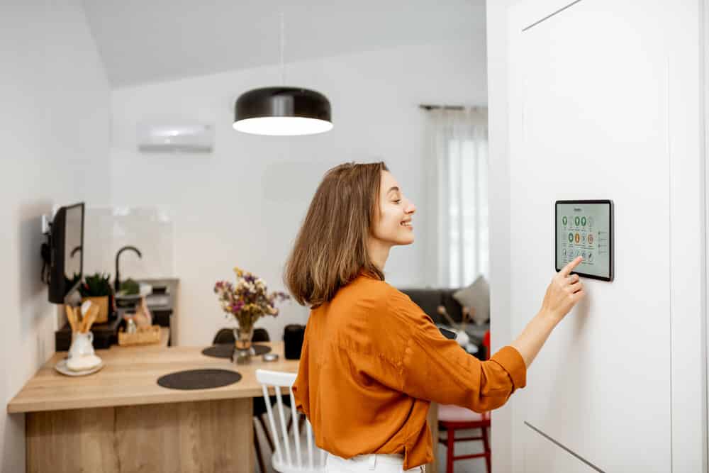 Young woman controlling home with a digital touch screen panel installed on the wall in the living room. Concept of a smart home and mobile application for managing smart devices at home. Example of 2025 remodeling trends