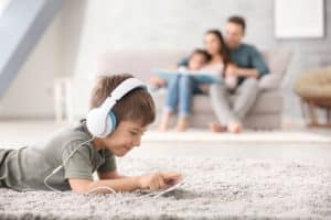 Cute little boy with tablet computer at home laying on the carpet with family behind him on the couch. Comfort is one of the pros of carpet compared to hardwood floors