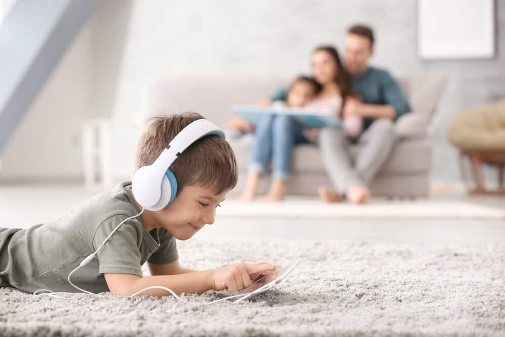 Cute little boy with tablet computer at home laying on the carpet with family behind him on the couch. Comfort is one of the pros of carpet compared to hardwood floors